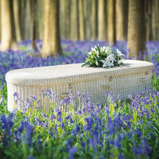 Handwoven natural wicker eco coffin adorned with a bouquet of white flowers, placed in a tranquil bluebell forest.