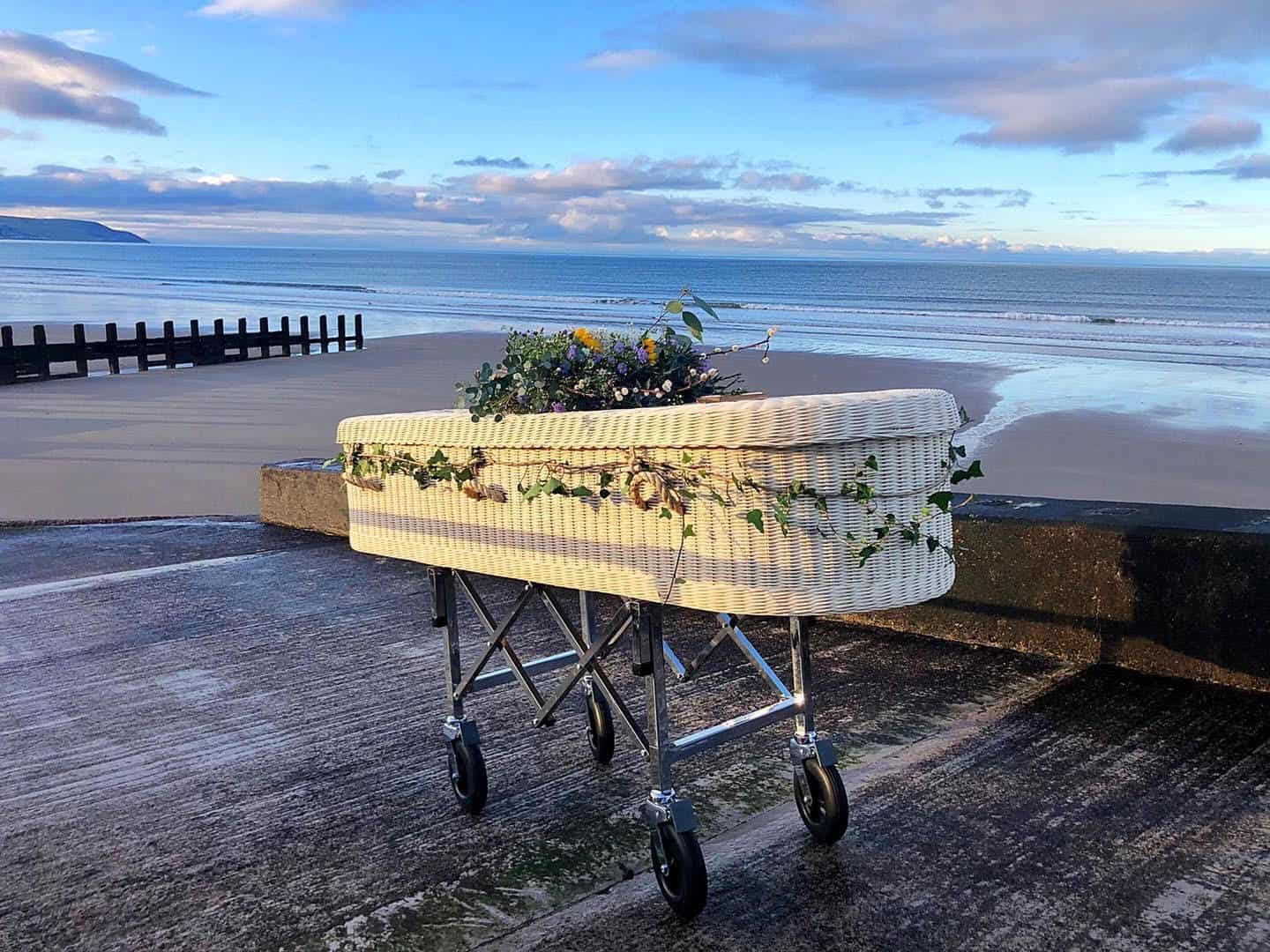 Sustainably crafted wicker coffin with floral garland decoration, positioned on a trolley overlooking a peaceful beach at low tide.