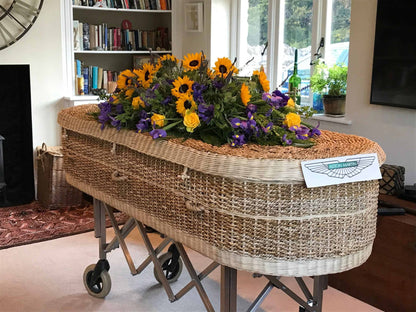 Artisanal woven coffin lavishly adorned with bright sunflowers, purple blooms, and yellow roses, presented in a cozy room with a view of a garden.