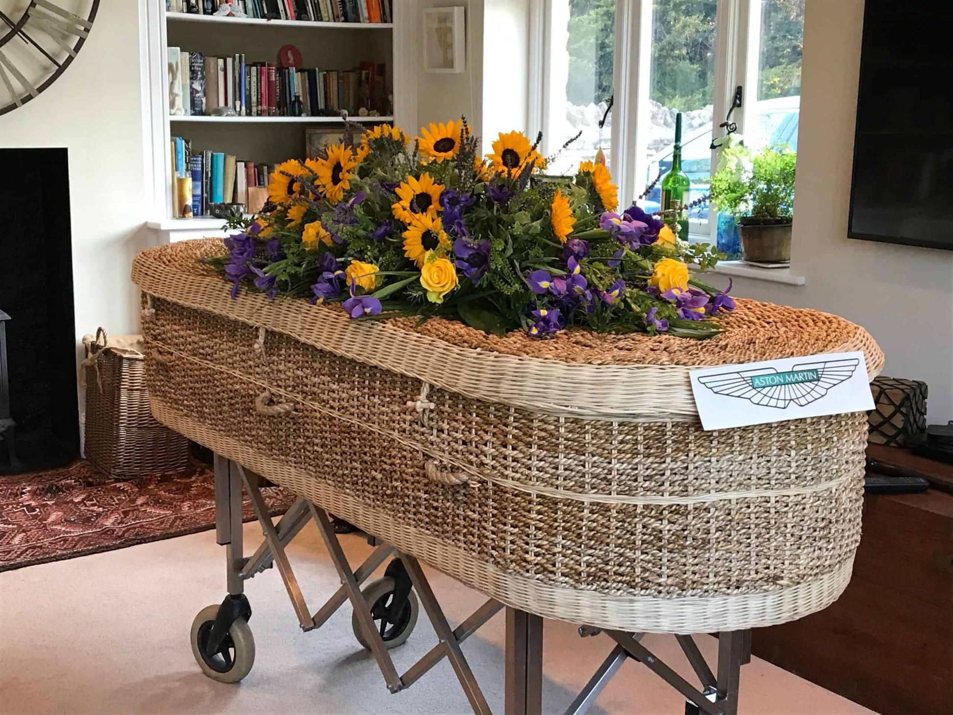 Elegantly crafted wicker coffin embellished with a colorful array of sunflowers and purple flowers, displayed on a trolley in a home library with a garden view.